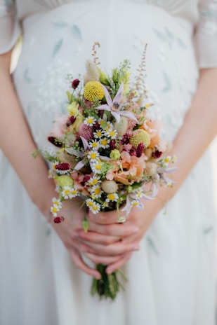 A person is holding a floral bouquet with a variety of small, colorful flowers including daisies, yellow billy balls, and pink blossoms. The hands gently clasp the stems, and the background shows a white dress with subtle embroidery.