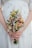 A person is holding a floral bouquet with a variety of small, colorful flowers including daisies, yellow billy balls, and pink blossoms. The hands gently clasp the stems, and the background shows a white dress with subtle embroidery.