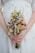 A person is holding a floral bouquet with a variety of small, colorful flowers including daisies, yellow billy balls, and pink blossoms. The hands gently clasp the stems, and the background shows a white dress with subtle embroidery.