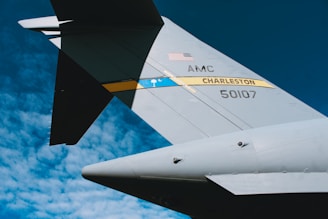 A close-up view of the tail section of a military aircraft with the text 'AMC CHARLESTON' and '50107' on it, featuring an American flag and a blue stripe with a symbol. The background consists of a clear blue sky with scattered clouds.