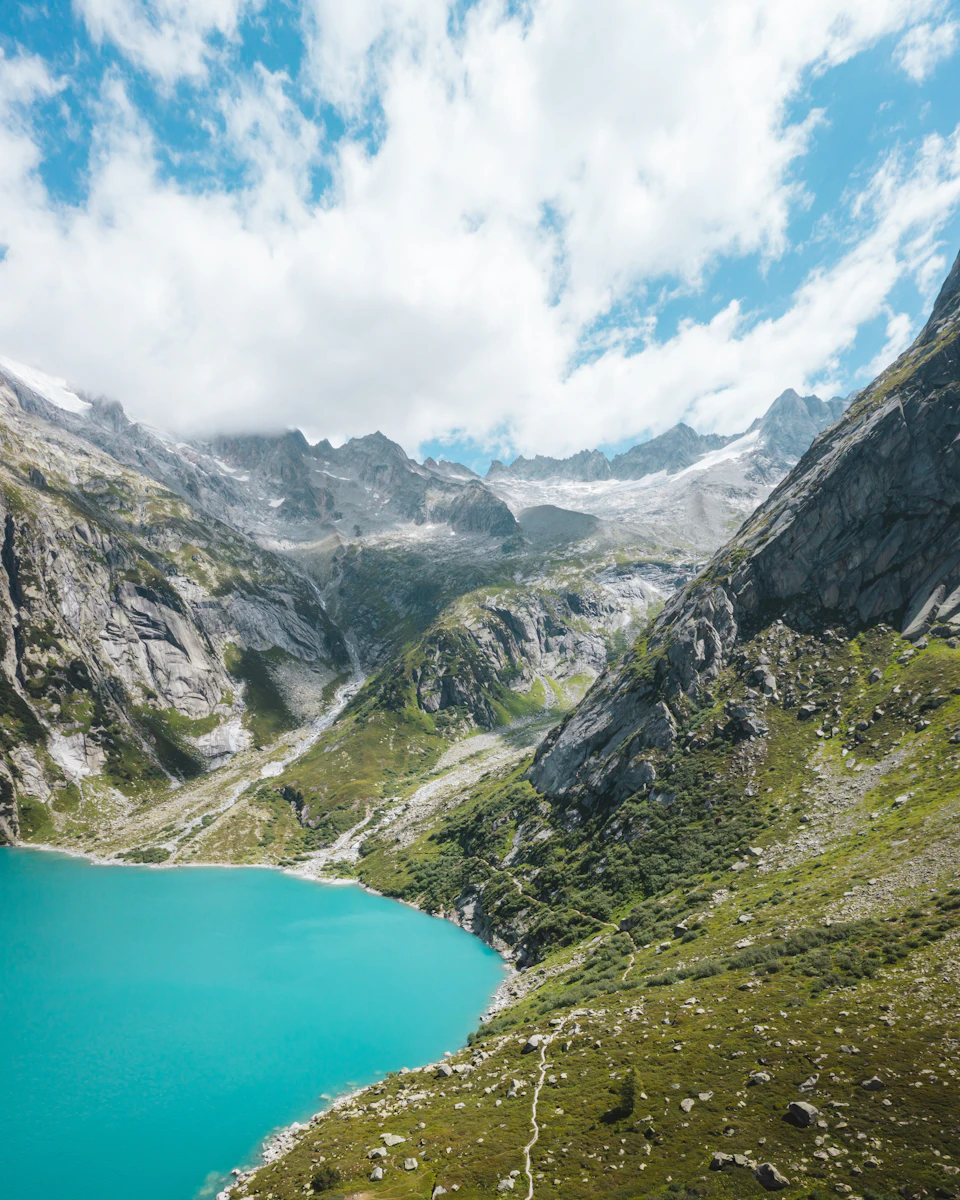 The image depicts a breathtaking view of a mountain range with a deep blue lake nestled at its base, surrounded by lush green vegetation and towering mountains. The sky above is a light blue canvas dotted with fluffy white clouds, creating a serene atmosphere. The perspective of the photo is from a high vantage point, providing an expansive view of the landscape below.