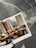 A magazine open to a page showing a cozy living room with a brown sectional sofa adorned with plush pillows, two round wooden side tables, and a large window revealing a snowy landscape. The magazine is placed on a textured stone surface with gray and white patterns.