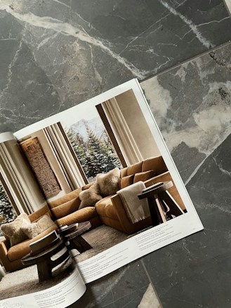 A magazine open to a page showing a cozy living room with a brown sectional sofa adorned with plush pillows, two round wooden side tables, and a large window revealing a snowy landscape. The magazine is placed on a textured stone surface with gray and white patterns.