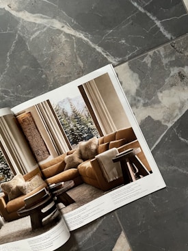 A magazine open to a page showing a cozy living room with a brown sectional sofa adorned with plush pillows, two round wooden side tables, and a large window revealing a snowy landscape. The magazine is placed on a textured stone surface with gray and white patterns.