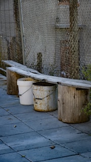 A makeshift bench is created by placing a long wooden plank over three buckets. One of the buckets is white and two are a rusty yellow color. The bench is situated next to a chain-link fence with a building in the background. The ground is covered in stone tiles with some scattered leaves.