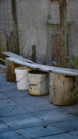 A makeshift bench is created by placing a long wooden plank over three buckets. One of the buckets is white and two are a rusty yellow color. The bench is situated next to a chain-link fence with a building in the background. The ground is covered in stone tiles with some scattered leaves.