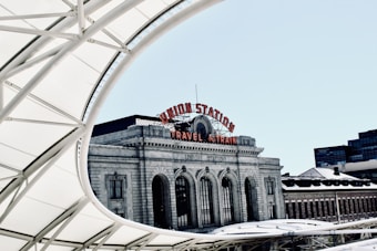 A historic train station building with a large, arched window facade and a sign reading 'Union Station' and 'Travel by Train' in red letters. The architecture is ornate and classical, featuring decorative stonework beneath a bright blue sky. Modern structural elements, like a white, curved metal and fabric canopy, frame the view.