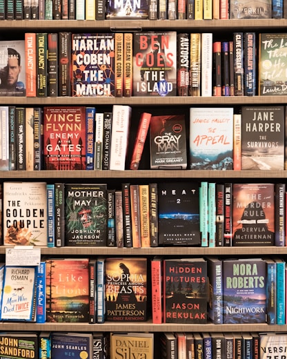A bookshelf filled with a variety of novels, each with colorful and distinct covers. The titles include works by authors such as Harlan Coben, Ben Coes, and Vince Flynn. The books are neatly arranged in multiple rows, showcasing different genres including thrillers and mysteries.