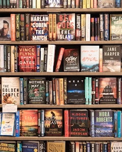 A bookshelf filled with a variety of novels, each with colorful and distinct covers. The titles include works by authors such as Harlan Coben, Ben Coes, and Vince Flynn. The books are neatly arranged in multiple rows, showcasing different genres including thrillers and mysteries.