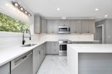 A modern kitchen with sleek gray cabinets and white countertops. The stainless steel appliances include a dishwasher and an oven with a microwave above it. A large window provides natural light, and recessed lighting lines the ceiling. The floor is fitted with white marble tiles.