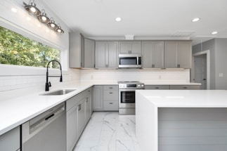 A modern kitchen with sleek gray cabinets and white countertops. The stainless steel appliances include a dishwasher and an oven with a microwave above it. A large window provides natural light, and recessed lighting lines the ceiling. The floor is fitted with white marble tiles.