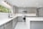 A modern kitchen with sleek gray cabinets and white countertops. The stainless steel appliances include a dishwasher and an oven with a microwave above it. A large window provides natural light, and recessed lighting lines the ceiling. The floor is fitted with white marble tiles.