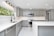 A modern kitchen with sleek gray cabinets and white countertops. The stainless steel appliances include a dishwasher and an oven with a microwave above it. A large window provides natural light, and recessed lighting lines the ceiling. The floor is fitted with white marble tiles.