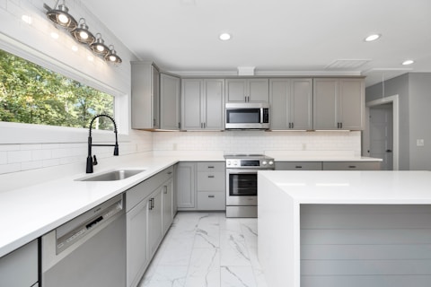 A modern kitchen with sleek gray cabinets and white countertops. The stainless steel appliances include a dishwasher and an oven with a microwave above it. A large window provides natural light, and recessed lighting lines the ceiling. The floor is fitted with white marble tiles.
