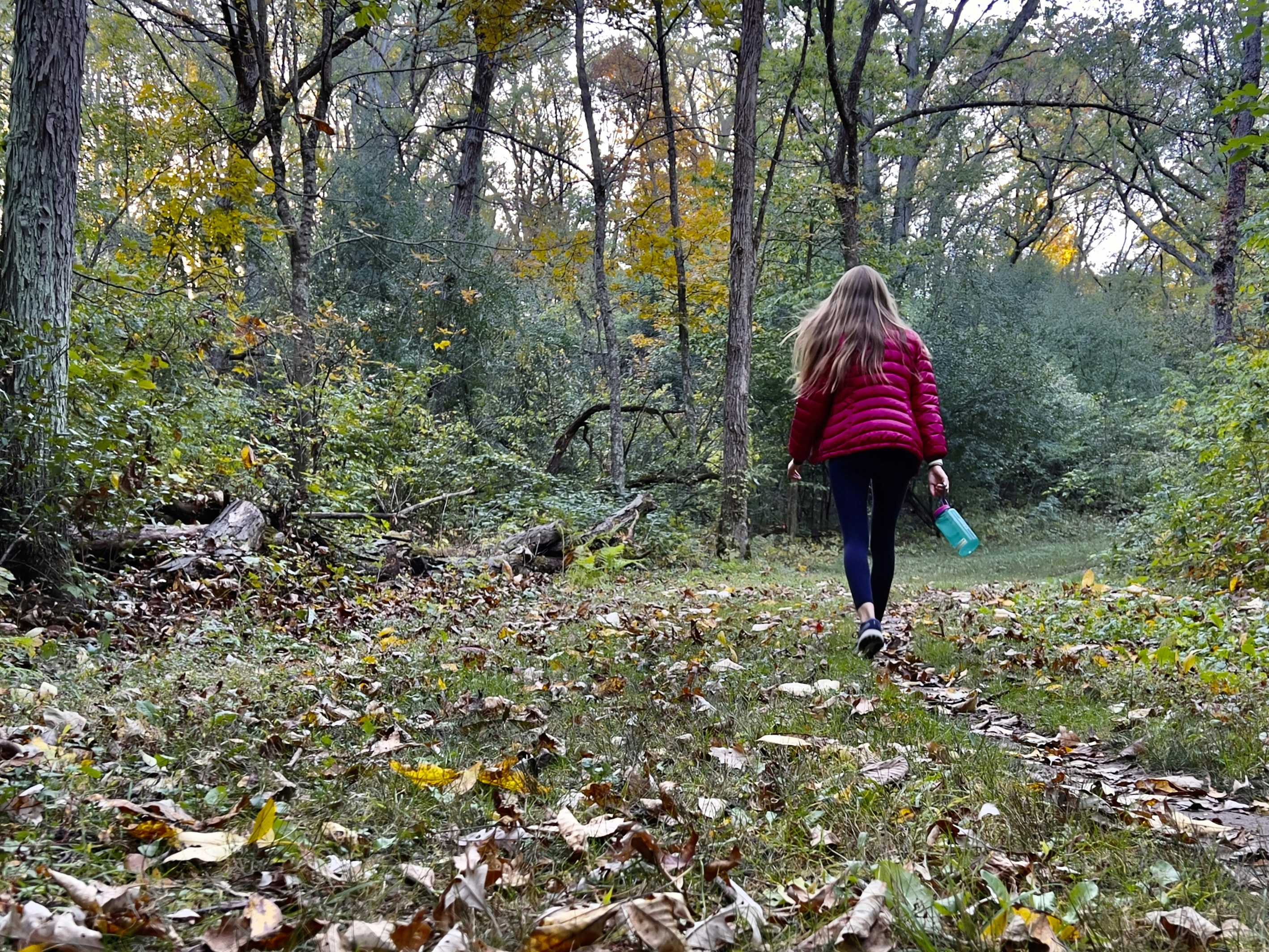 Frau in roter Jacke spaziert auf einem laubbedeckten Pfad in einen herbstlichen Wald hinein
