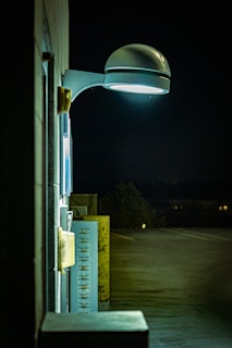 A nighttime scene featuring a wall-mounted, modern street lamp illuminating a section of a building and adjacent utility panels. The light casts a cool white glow, creating shadows and highlighting textures on the wall. The background reveals an empty parking lot with minimal lighting.