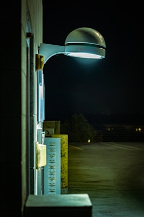A nighttime scene featuring a wall-mounted, modern street lamp illuminating a section of a building and adjacent utility panels. The light casts a cool white glow, creating shadows and highlighting textures on the wall. The background reveals an empty parking lot with minimal lighting.