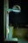 A nighttime scene featuring a wall-mounted, modern street lamp illuminating a section of a building and adjacent utility panels. The light casts a cool white glow, creating shadows and highlighting textures on the wall. The background reveals an empty parking lot with minimal lighting.
