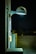 A nighttime scene featuring a wall-mounted, modern street lamp illuminating a section of a building and adjacent utility panels. The light casts a cool white glow, creating shadows and highlighting textures on the wall. The background reveals an empty parking lot with minimal lighting.