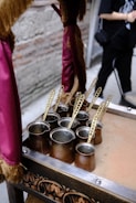 Several traditional copper coffee pots with long, ornate handles are placed on a decorated table or tray. The background includes a blurred figure and a wall with exposed brick. A piece of maroon fabric with gold fringe hangs nearby, suggesting an elegant or vintage setting.