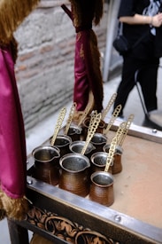 Several traditional copper coffee pots with long, ornate handles are placed on a decorated table or tray. The background includes a blurred figure and a wall with exposed brick. A piece of maroon fabric with gold fringe hangs nearby, suggesting an elegant or vintage setting.
