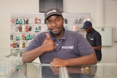 A man wearing a cap and a shirt with 'ProCell' logo, giving a thumbs-up while leaning on a glass display counter. The background shows another individual and shelves filled with various mobile phone accessories.