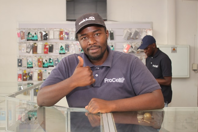 A man wearing a cap and a shirt with 'ProCell' logo, giving a thumbs-up while leaning on a glass display counter. The background shows another individual and shelves filled with various mobile phone accessories.