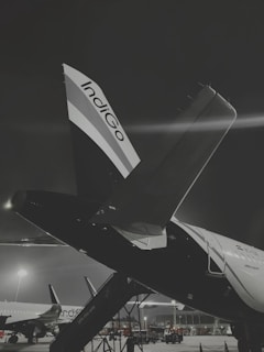 A nighttime scene at an airport, prominently featuring an airplane with the company logo Indigo on its tail. The aircraft is stationed on the tarmac with visible boarding stairs. Other planes are visible in the background, under the glow of bright airport lights. The overall scene conveys a sense of calm yet busy atmosphere at a well-lit airport.