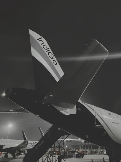 A nighttime scene at an airport, prominently featuring an airplane with the company logo Indigo on its tail. The aircraft is stationed on the tarmac with visible boarding stairs. Other planes are visible in the background, under the glow of bright airport lights. The overall scene conveys a sense of calm yet busy atmosphere at a well-lit airport.