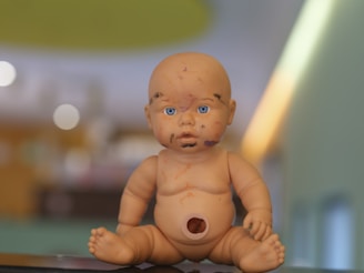 A baby doll with blue eyes is sitting on a surface. The doll's face and body have various markings or scribbles in different colors. The background is blurred, displaying warm tones and abstract shapes.