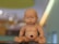 A baby doll with blue eyes is sitting on a surface. The doll's face and body have various markings or scribbles in different colors. The background is blurred, displaying warm tones and abstract shapes.