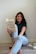 A woman is sitting on the floor against a plain wall, holding and smiling at a book titled 'Atomic Habits'. She is wearing a dark green shirt and light blue jeans. There are additional books and a small red cup on the floor next to her.