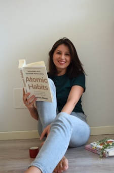 A woman is sitting on the floor against a plain wall, holding and smiling at a book titled 'Atomic Habits'. She is wearing a dark green shirt and light blue jeans. There are additional books and a small red cup on the floor next to her.