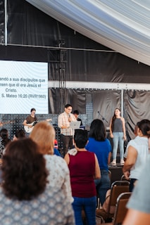A group of people gathered in what appears to be a religious service or event. There is a stage with musicians and speakers while attendees listen. A large screen displays a Bible verse in Spanish.