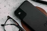 A sleek black power bank with an LED display indicating 88% charge lies on a brown leather surface. Nearby are a pair of black-rimmed eyeglasses and a black pen, all against a textured light-colored background.
