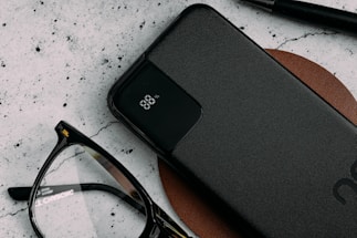 A sleek black power bank with an LED display indicating 88% charge lies on a brown leather surface. Nearby are a pair of black-rimmed eyeglasses and a black pen, all against a textured light-colored background.