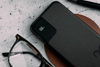 A sleek black power bank with an LED display indicating 88% charge lies on a brown leather surface. Nearby are a pair of black-rimmed eyeglasses and a black pen, all against a textured light-colored background.