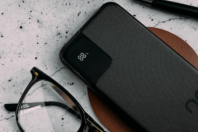 A sleek black power bank with an LED display indicating 88% charge lies on a brown leather surface. Nearby are a pair of black-rimmed eyeglasses and a black pen, all against a textured light-colored background.