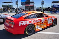 A brightly colored race car with a red and orange design featuring the text 'Race for a Cure' and the number 16. The car is parked on a track with various sponsorship logos visible, including Goodyear and Monster Energy. In the background, there are people walking and equipment related to the racing event.