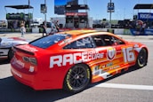 A brightly colored race car with a red and orange design featuring the text 'Race for a Cure' and the number 16. The car is parked on a track with various sponsorship logos visible, including Goodyear and Monster Energy. In the background, there are people walking and equipment related to the racing event.