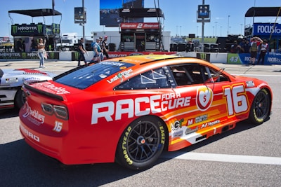 A brightly colored race car with a red and orange design featuring the text 'Race for a Cure' and the number 16. The car is parked on a track with various sponsorship logos visible, including Goodyear and Monster Energy. In the background, there are people walking and equipment related to the racing event.
