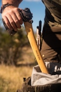 A person is holding the handle of an axe, which has a protective cover on its head. The axe is branded with the logo 'Hults Bruk.' The tool is being placed into a backpack, and the setting appears to be outdoors with a blurred background of grass and foliage.