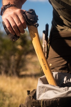 A person is holding the handle of an axe, which has a protective cover on its head. The axe is branded with the logo 'Hults Bruk.' The tool is being placed into a backpack, and the setting appears to be outdoors with a blurred background of grass and foliage.