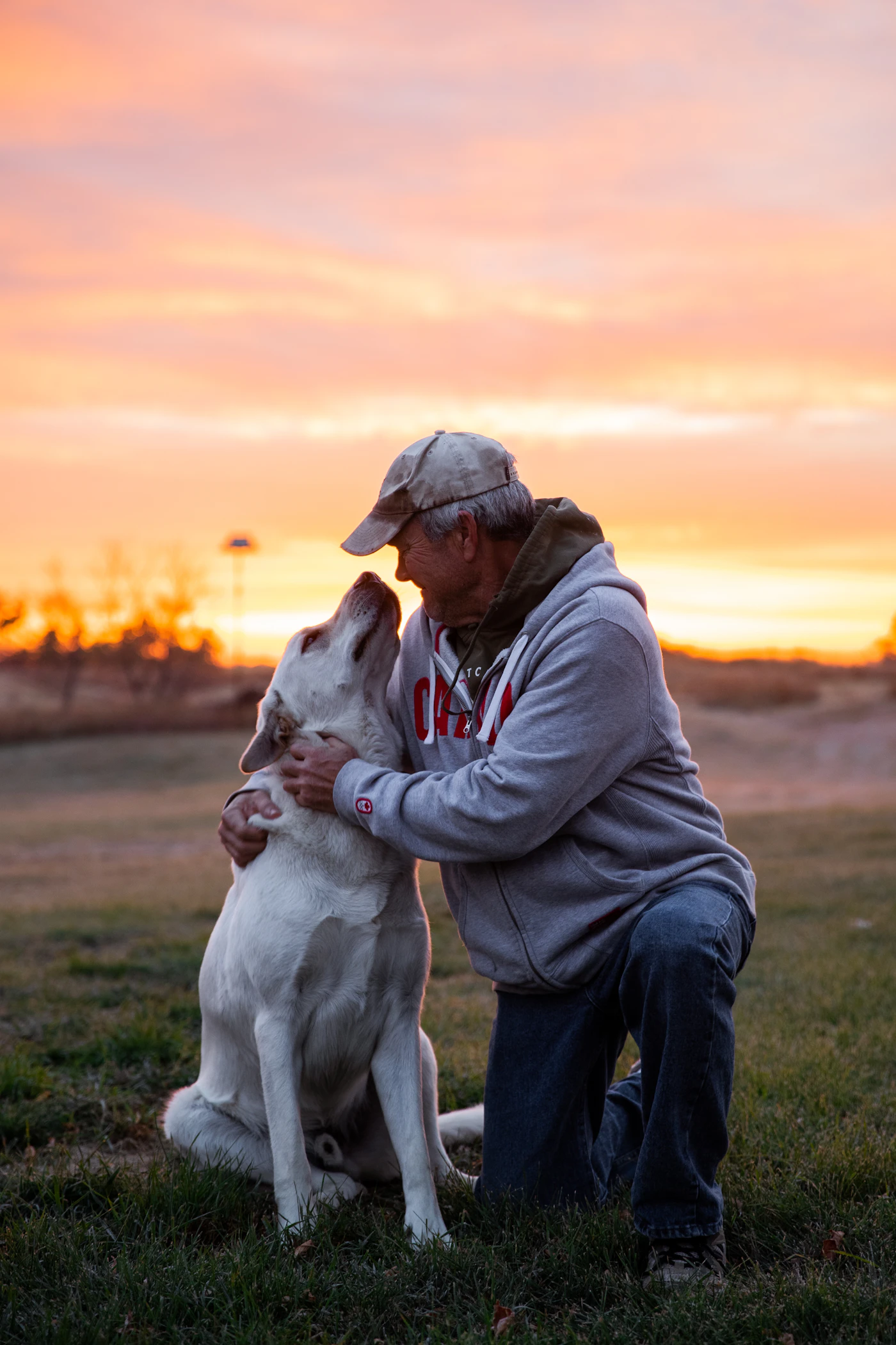 An older man kneeling face to face with his white Labrador in an open field as the sky glows orange at sunset