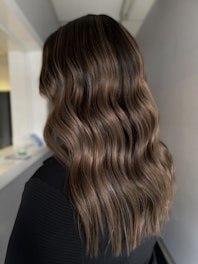 Long, wavy brown hair cascading down the back of a person with soft highlights. The background includes a blurred indoor setting, possibly a hallway.