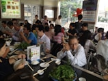 A bustling restaurant with many people seated at tables, engaging in conversation and eating. There is a variety of dishes on the tables, including leafy greens and rice. The room is bright, with natural light coming through large windows.