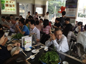 A bustling restaurant with many people seated at tables, engaging in conversation and eating. There is a variety of dishes on the tables, including leafy greens and rice. The room is bright, with natural light coming through large windows.