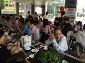 A bustling restaurant with many people seated at tables, engaging in conversation and eating. There is a variety of dishes on the tables, including leafy greens and rice. The room is bright, with natural light coming through large windows.