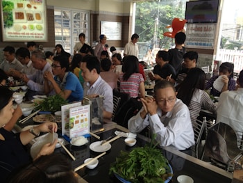 A bustling restaurant with many people seated at tables, engaging in conversation and eating. There is a variety of dishes on the tables, including leafy greens and rice. The room is bright, with natural light coming through large windows.