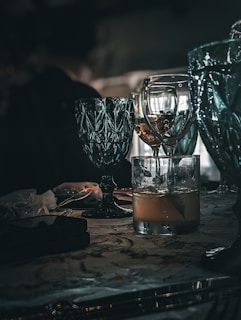 A collection of elegant glassware is placed on a table, featuring a mix of turquoise and clear glasses. The scene is dimly lit, creating reflections and shadows that enhance the luxurious feel. The table surface has a patterned design and holds additional items, including a small black object and a translucent bag.